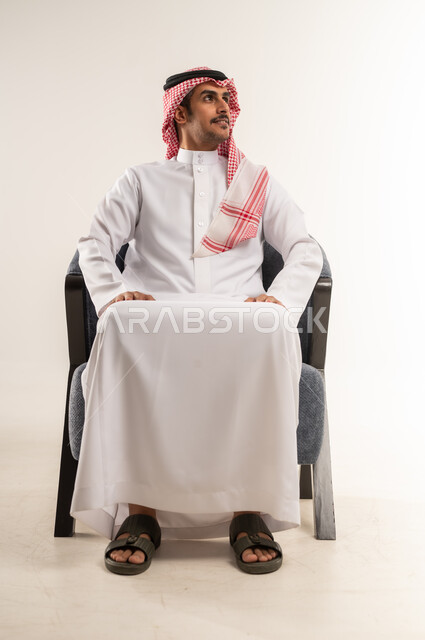 التقاط الصور الشخصية للوثائق الرسمية، الاهتمام بالمظهر الخارجي والهندام، رجل عربي خليجي بحريني يرتدي الثوب التقليدي والشماغ يجلس على كرسي مريح بتعابير الثقة بالنفس، النظر إلى مكان ما بإيماءات الاندماج، مفهوم الأناقة والشياكة، الجسم بالطول الكامل، خلفية بيضاء