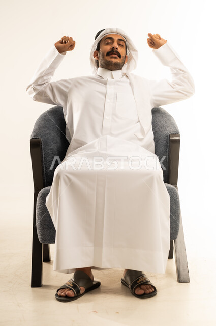 الإحساس بالكسل والخمول، بورتريه لرجل عربي خليجي قطري يرتدي الثوب التقليدي والغترة يجلس على كرسي مريح يقوم بتمديد جسمه ورفع ذراعيه للاعلى بإيماءات النعاس والتثاوب والارهاق, الشعور بالتعب والحاجة الى النوم والراحة, خلفية بيضاء