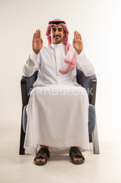 مفهوم الأناقة والشياكة، الاهتمام بالمظهر الخارجي والهندام، رجل عربي خليجي بحريني يرتدي الثوب التقليدي والشماغ يجلس على كرسي مريح يفتح كفيه ويرفعهما للأعلى، النظر للكاميرا بإيماءات البشاشة، التعبير بحركات اليدين ولغة الإشارة، الجسم بالطول الكامل، خلفية بيضاء