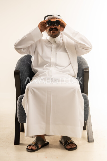 التخطيط ووضع الأهداف المستقبلية، استخدام المنظار ثنائي العينين لمشاهدة الأشياء البعيدة، توضيح الرؤية وتقريبها، بورتريه لرجل عربي خليجي قطري يرتدي الثوب التقليدي والغترة يجلس على كرسي ويضع تلسكوب العدسات المقربة على عينيه، خلفية بيضاء