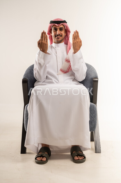التعبير بحركات اليدين ولغة الإشارة، مفهوم الأناقة والشياكة، الاهتمام بالمظهر الخارجي والهندام، رجل عربي خليجي بحريني يرتدي الثوب التقليدي والشماغ يجلس على كرسي مريح يفتح كفيه ويرفعهما للأعلى، النظر للكاميرا بإيماءات البشاشة، الجسم بالطول الكامل، خلفية بيضاء