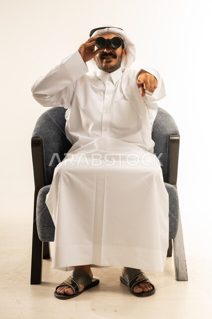 استخدام المنظار ثنائي العينين، التخطيط ووضع الأهداف المستقبلية، بورتريه لرجل عربي خليجي قطري يرتدي الثوب التقليدي والغترة يجلس على كرسي ويضع تلسكوب ذو العدسات المقربة على عينيه، الإشارة إلى الكاميرا باصبع السبابة بشكل مباشر، خلفية بيضاء