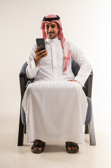 Communicating with individuals via mobile phone, using modern techniques and devices and technology, browsing applications and websites, chatting and conversations with friends, portrait of a Bahraini Gulf Arab man wearing a traditional dress and shemagh sitting on a comfortable chair, full body length, white background