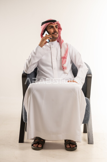 Making phone calls via mobile phone, using modern techniques and devices and technology, communicating with individuals via the Internet, chatting and conversations with friends, portrait of a Bahraini Gulf Arab man wearing a traditional thobe and shemagh sitting on a comfortable chair, full body length, white background
