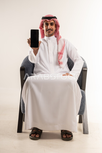 رفع الهاتف المحمول بشاشة سوداء فارغة، الإعلانات الترويجية والتسويقة، استخدام التقنيات الحديثة والتكنولوجيا, النظر للكاميرا بإيماءات البشاشة، بورتريه لرجل عربي خليجي بحريني يرتدي الثوب التقليدي والشماغ يجلس على كرسي مريح، الطول الكامل للجسم، خلفية بيضاء