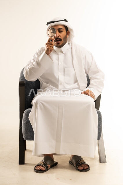 مضاعفة الأحجام ومحاولة إيجاد الأشياء الصغيرة، بورتريه لرجل عربي خليجي قطري يرتدي الثوب التقليدي والغترة يجلس على كرسي يحاول اكتشاف شيء ما باستخدام العدسة المكبرة، إيماءات الاندماج والتركيز، تكبير وتوضيح الرؤية، عمليات البحث والتدقيق، خلفية بيضاء