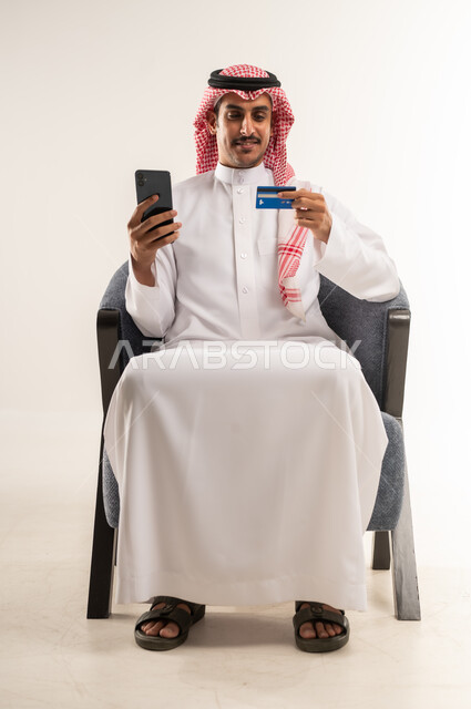 Electronic payment based on bank card, online shopping concept, shopping and purchasing operations via internet applications, use of modern techniques and technology, portrait of a Bahraini Gulf Arab man wearing traditional thobe and shemagh sitting on a chair, full body length, white background