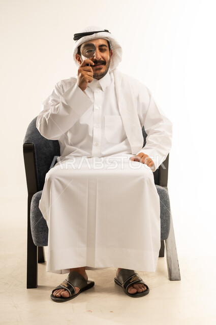 عمليات البحث والتدقيق، مضاعفة الأحجام ومحاولة إيجاد الأشياء الصغيرة، بورتريه لرجل عربي خليجي قطري يرتدي الثوب التقليدي والغترة يجلس على كرسي يحاول اكتشاف شيء ما باستخدام العدسة المكبرة، إيماءات الاندماج والسرور، تكبير وتوضيح الرؤية، خلفية بيضاء