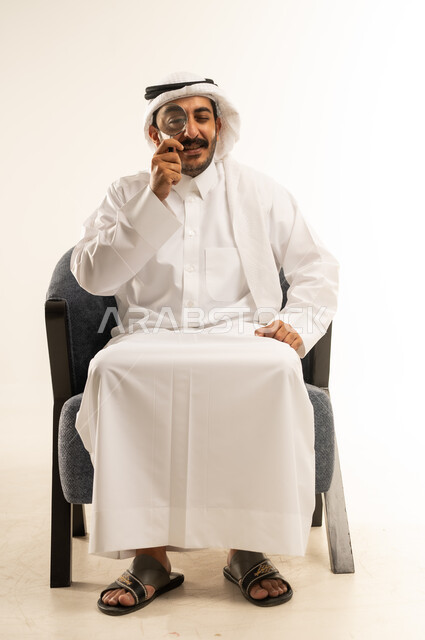 تكبير وتوضيح الرؤية، عمليات البحث والتدقيق، مضاعفة الأحجام ومحاولة إيجاد الأشياء الصغيرة، بورتريه لرجل عربي خليجي قطري يرتدي الثوب التقليدي والغترة يجلس على كرسي يحاول اكتشاف شيء ما باستخدام العدسة المكبرة، إيماءات الاندماج والسرور، خلفية بيضاء