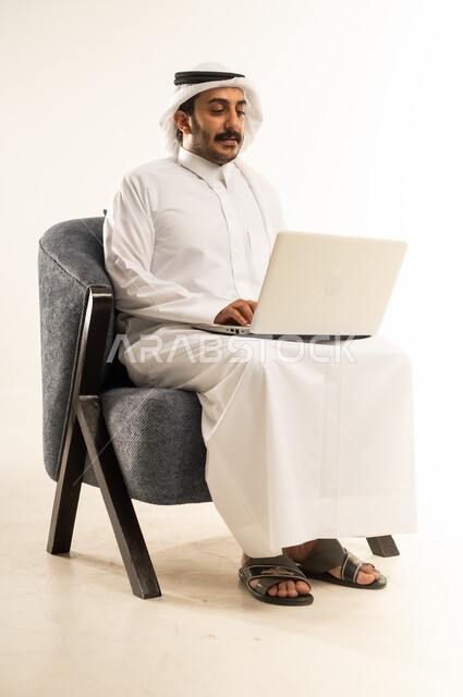 مفهوم التوظيف أونلاين، تصفح مواقع التواصل الاجتماعي بواسطة الحاسب المحمول، العمل عن بعد عبر الانترنت، دمج التكنولوجيا في الحياة العملية, بورتريه لرجل عربي خليجي قطري يرتدي الثوب التقليدي والغترة يجلس على كرسي مريح يقوم باستخدام جهاز اللابتوب، خلفية بيضاء