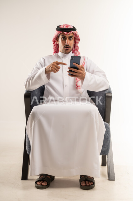 الإشارة إلى الهاتف المحمول باصبع السبابة بإيماءات المفاجأة والدهشة، استخدام التقنيات والأجهزة الحديثة والتكنولوجيا, رؤية اخبار صادمة وغير متوقعة، بورتريه لرجل عربي خليجي بحريني يرتدي الثوب التقليدي والشماغ يجلس على كرسي مريح، الطول الكامل للجسم، خلفية بيضاء