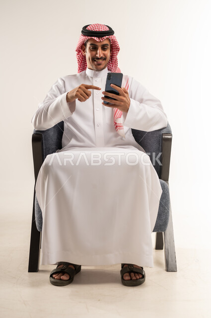 رؤية وسماع أخبار سعيدة ومفرحة، الإشارة إلى الهاتف المحمول باصبع السبابة بإيماءات السعادة والبشاشة، استخدام التقنيات والأجهزة الحديثة والتكنولوجيا, بورتريه لرجل عربي خليجي بحريني يرتدي الثوب التقليدي والشماغ يجلس على كرسي مريح، الطول الكامل للجسم، خلفية بيضاء