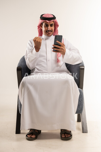 رفع قبضة اليد نحو الأعلى بتعابير الحماس، النظر إلى الهاتف المحمول بإيماءات السعادة والبشاشة، استخدام التقنيات والأجهزة الحديثة والتكنولوجيا, بورتريه لرجل عربي خليجي بحريني يرتدي الثوب التقليدي والشماغ يجلس على كرسي مريح، الطول الكامل للجسم، خلفية بيضاء