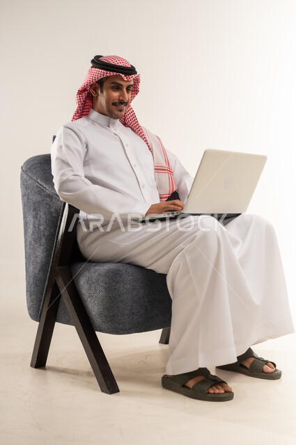 العمل عن بعد عبر الانترنت، إنجاز المهام بسرعة واحترافية بالاعتماد على الحاسوب المحمول، مفهوم التوظيف أونلاين، استخدام التقنيات الحديثة والتكنولوجيا, بورتريه لرجل عربي خليجي بحريني يرتدي الثوب التقليدي والشماغ يجلس على كرسي، الطول الكامل للجسم، خلفية بيضاء