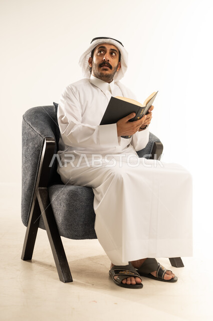 قراءة الكتب والقصص والروايات، تنمية المهارات وتوسيع المعارف والأفكار، بورتريه لرجل عربي خليجي قطري يرتدي الثوب التقليدي والغترة يجلس على كرسي مريح يمسك بيديه كتاب، النظر الى مكان ما بإيماءات التفكير والتمعن، زيادة الذخيرة الفكرية، خلفية بيضاء