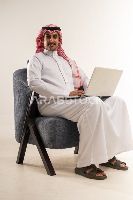 إنجاز المهام بسرعة واحترافية بالاعتماد على الحاسوب المحمول، مفهوم التوظيف أونلاين، العمل عن بعد عبر الانترنت، استخدام التقنيات الحديثة والتكنولوجيا, بورتريه لرجل عربي خليجي بحريني يرتدي الثوب التقليدي والشماغ يجلس على كرسي، الطول الكامل للجسم، خلفية بيضاء