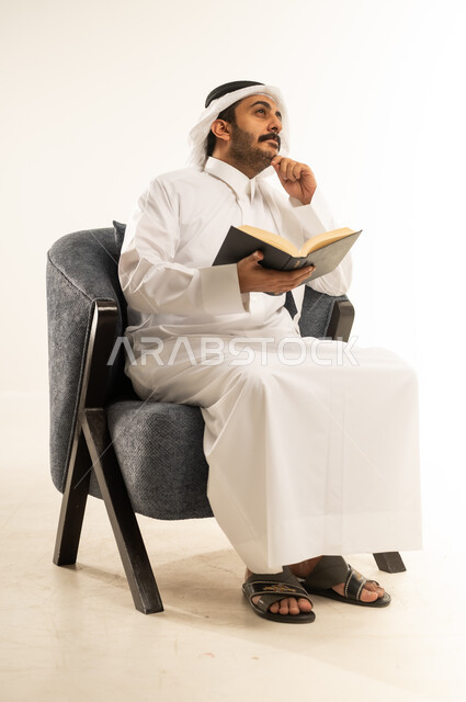 زيادة الذخيرة الفكرية، قراءة الكتب والقصص والروايات، تنمية المهارات وتوسيع المعارف والأفكار، بورتريه لرجل عربي خليجي قطري يرتدي الثوب التقليدي والغترة يجلس على كرسي مريح يمسك بيديه كتاب ويضع يده تحت ذقنه، النظر الى مكان ما بإيماءات التفكير والتمعن، خلفية بيضاء
