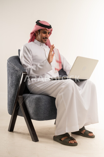 Holding remote business meetings via the Internet, making video calls using a laptop, the concept of online recruitment, integrating technology with administrative work, portrait of a Bahraini Gulf Arab man wearing a traditional thobe and shemagh sitting on a chair, full body length, white background