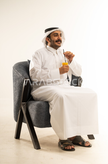 الجلوس ومحاولة الراحة والاستمتاع بأوقات الفراغ، بورتريه لشاب عربي خليجي قطري يرتدي الثوب التقليدي والغترة يجلس على كرسي مريح يمسك بيده كوب من المشروب الصيفي المفضل، الحصول على كأس من العصير البارد والمنعش، خلفية بيضاء