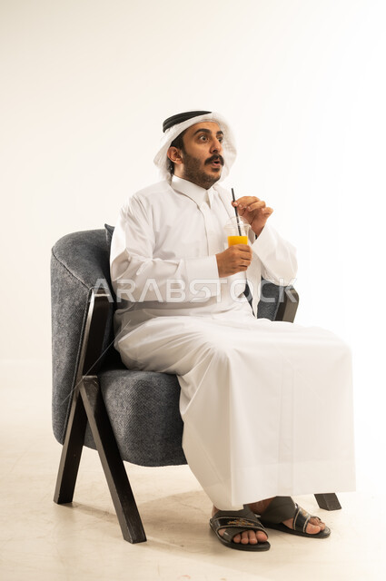 ايماءات المفاجأة والدهشة، بورتريه لشاب عربي خليجي قطري يرتدي الثوب التقليدي والغترة يجلس على كرسي مريح يمسك بيده كوب من المشروب الصيفي المفضل، الحصول على كأس من العصير البارد والمنعش، الجلوس ومحاولة الراحة والاستمتاع بأوقات الفراغ، خلفية بيضاء