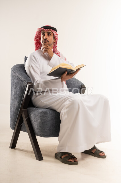 تنمية المهارات وتوسيع المعارف والأفكار، زيادة الذخيرة الفكرية، قضاء أوقات الفراغ بشكل مفيد، الاستمتاع بقراءة الكتب والقصص والروايات، بورتريه لشاب عربي خليجي بحريني يرتدي الثوب التقليدي والشماغ يجلس على كرسي ويمسك بيديه كتاب، الطول الكامل للجسم، خلفية بيضاء