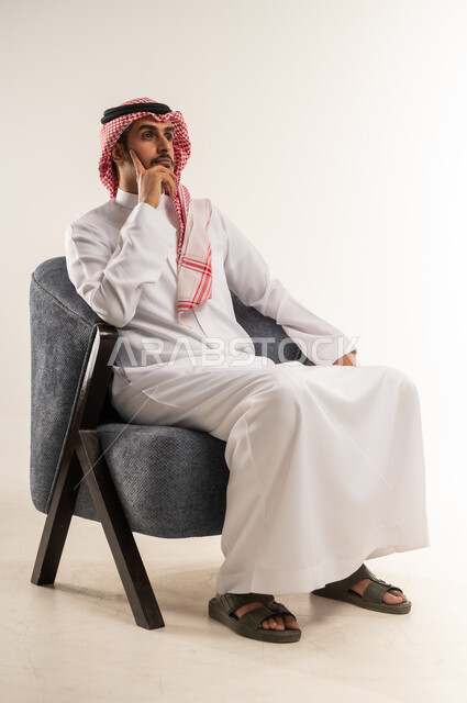 وضع اليد على الخد وشرود الذهن، التفكير بشكل عميق، محاولة إيجاد حل للمشكلات الصعبة، بورتريه لشاب عربي خليجي بحريني يرتدي الثوب التقليدي والشماغ يجلس على كرسي وينظر إلى مكان بإيماءات الاندماج، الطول الكامل للجسم، خلفية بيضاء