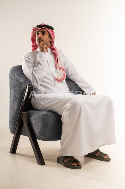 التفكير بشكل عميق، وضع اليد على الخد وشرود الذهن، محاولة إيجاد حل للمشكلات الصعبة، بورتريه لشاب عربي خليجي بحريني يرتدي الثوب التقليدي والشماغ يجلس على كرسي وينظر إلى مكان بإيماءات الاندماج، الطول الكامل للجسم، خلفية بيضاء