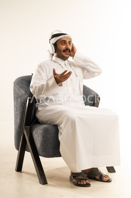دمج التقنيات والأجهزة الحديثة والتكنولوجيا بالحياة اليومية، الاستماع إلى الموسيقا والبودكاست، استخدام تقنية الهيدفون، بورتريه لرجل عربي خليجي قطري يرتدي الثوب التقليدي والغترة يجلس على كرسي مريح ويضع سماعة الأذنين، خلفية بيضاء