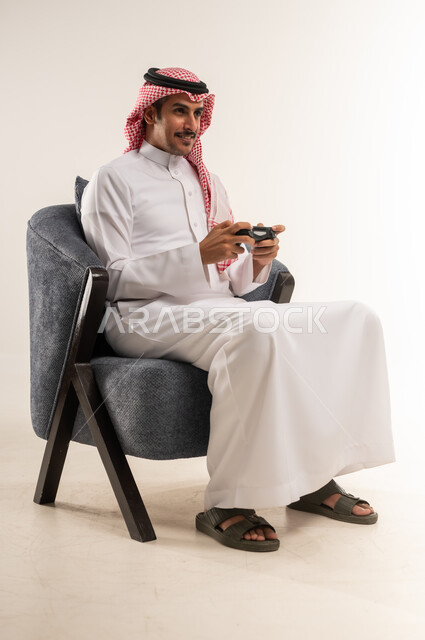 الاستمتاع بألعاب الفيديو المسلية، الحماس والتفاعل والاندماج مع اللعب، الاستفادة من التقنيات الحديثة والتكنولوجيا في أوقات الفراغ، بورتريه لشاب عربي خليجي بحريني يرتدي الثوب التقليدي والشماغ يجلس على كرسي ويمسك القبضة بيديه، الطول الكامل للجسم، خلفية بيضاء