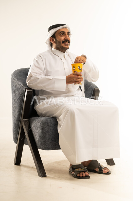 Sitting and watching movies in free time, gestures of integration and concentration with the movie, enjoying eating popcorn, portrait of a young Arab Gulf Qatari man wearing traditional thobe and ghutra sitting on a comfortable chair and holding a box of delicious popcorn in his hand, white background