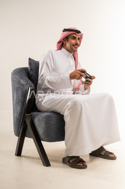 الحماس والتفاعل والاندماج مع اللعب، الاستمتاع بألعاب الفيديو المسلية، استخدام التقنيات الحديثة والتكنولوجيا في أوقات الفراغ، بورتريه لشاب عربي خليجي بحريني يرتدي الثوب التقليدي والشماغ يجلس على كرسي ويمسك القبضة بيديه، الطول الكامل للجسم، خلفية بيضاء