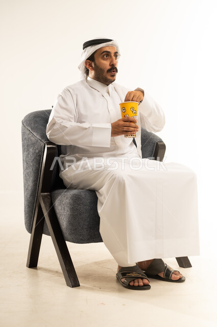 الاستمتاع بتناول الفشار، بورتريه لشاب عربي خليجي قطري يرتدي الثوب التقليدي والغترة يجلس على كرسي مريح ويمسك بيده علبة من البوب كورن اللذيذ ينظر الى مكان ما بتعابير الدهشة والذهول، الجلوس ومشاهدة الأفلام في أوقات الفراغ، خلفية بيضاء