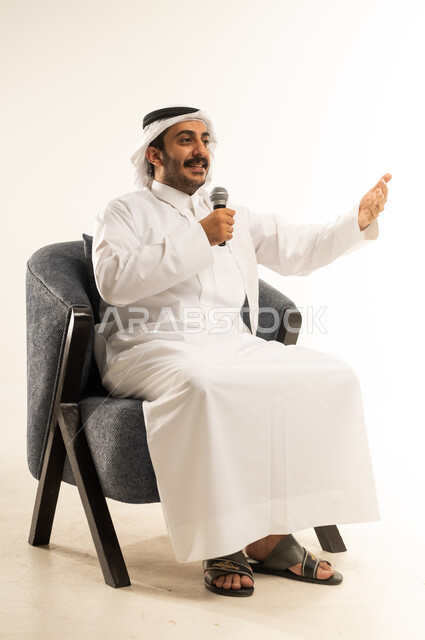 تنمية المواهب الصوتية والغنائية، بورتريه لرجل عربي خليجي قطري مبتسم يرتدي الثوب التقليدي والغترة يجلس على كرسي مريح يقوم بتسجيل مقطع بصوتي باستخدام مكبر الصوت، ايماءات الاستمتاع والتفاعل والاندماج مع الانغام الموسيقية ، خلفية بيضاء