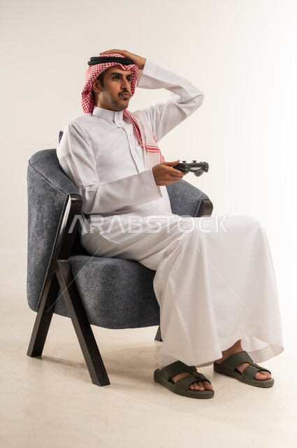 دمج التقنيات الحديثة والتكنولوجيا بالحياة اليومية، الحماس والاندماج مع اللعب، قضاء أوقات ممتعة بالتفاعل مع ألعاب الفيديو المسلية، بورتريه لشاب عربي خليجي بحريني يرتدي الثوب التقليدي والشماغ يجلس على كرسي ويمسك القبضة بيديه، الطول الكامل للجسم، خلفية بيضاء