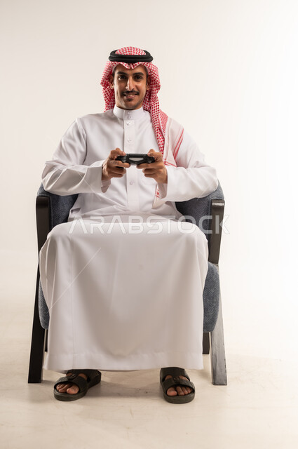 استخدام التقنيات الحديثة والتكنولوجيا في أوقات الفراغ، الحماس والتفاعل والاندماج مع اللعب، الاستمتاع بألعاب الفيديو المسلية، بورتريه لشاب عربي خليجي بحريني يرتدي الثوب التقليدي والشماغ يجلس على كرسي ويمسك القبضة بيديه، الطول الكامل للجسم، خلفية بيضاء