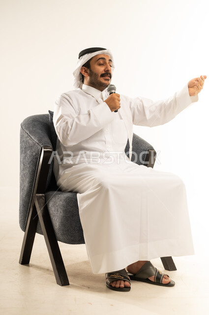ايماءات الاستمتاع والتفاعل والاندماج مع الانغام الموسيقية ، تنمية المواهب الصوتية والغنائية، بورتريه لرجل عربي خليجي قطري مبتسم يرتدي الثوب التقليدي والغترة يجلس على كرسي مريح يقوم بتسجيل مقطع بصوتي باستخدام مكبر الصوت ، خلفية بيضاء