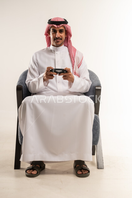 اللعب والاستمتاع بأوقات الفراغ، التفاعل مع ألعاب الفيديو بإيماءات الحماس والاندماج، دمج التقنيات الحديثة والتكنولوجيا بالحياة اليومية، بورتريه لشاب عربي خليجي بحريني يرتدي الثوب التقليدي والشماغ يجلس على كرسي ويمسك القبضة بيديه، الطول الكامل للجسم، خلفية بيضاء