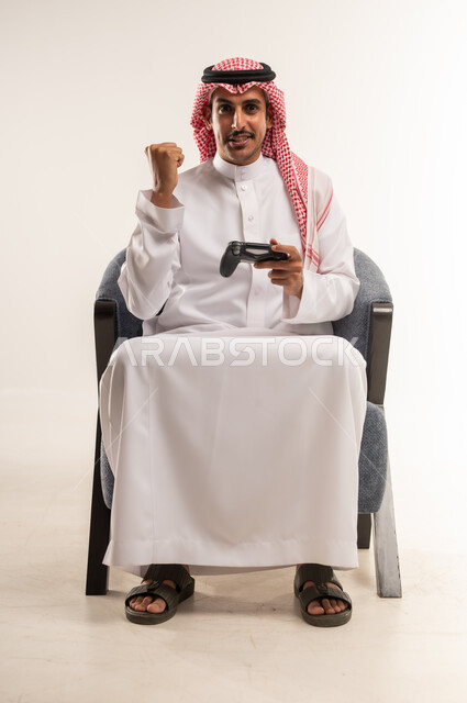 إيماءات الحماس والاندماج مع اللعب، قضاء أوقات ممتعة بالتفاعل مع ألعاب الفيديو، دمج التقنيات الحديثة والتكنولوجيا بالحياة اليومية،  بورتريه لشاب عربي خليجي بحريني يرتدي الثوب التقليدي والشماغ يجلس على كرسي ويمسك القبضة بيديه، الطول الكامل للجسم، خلفية بيضاء
