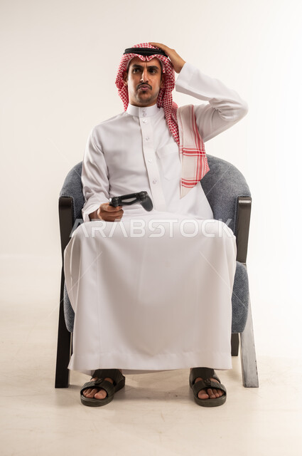 الإحباط والحزن على الخسارة في اللعب، التفاعل والاندماج مع ألعاب الفيديو، دمج التقنيات الحديثة والتكنولوجيا بالحياة اليومية، بورتريه لشاب عربي خليجي بحريني يرتدي الثوب التقليدي والشماغ يجلس على كرسي ويمسك قبضة اللعب بيديه، الطول الكامل للجسم، خلفية بيضاء