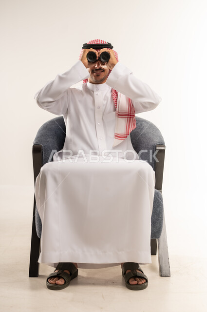 التخطيط ووضع الأهداف المستقبلية، استخدام المنظار ثنائي العينين لمشاهدة الأشياء البعيدة، توضيح الرؤية وتقريبها، بورتريه لشاب عربي خليجي بحريني يرتدي الثوب التقليدي والشماغ يجلس على كرسي ويضع تلسكوب العدسات المقربة على عينيه، الطول الكامل للجسم، خلفية بيضاء