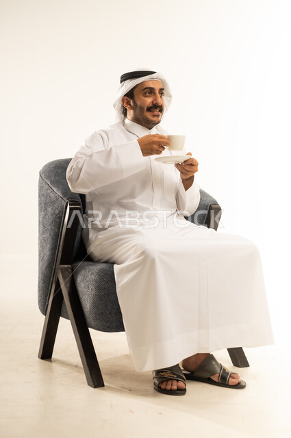 الاستمتاع بكوب من القهوة الساخنة، بورتريه لرجل عربي خليجي قطري يرتدي الثوب التقليدي والغترة يجلس على كرسي ويمسك بيده فنجان من مشروبه المفضل، محاولة الحصول على القليل من الراحة، الاسترخاء وتناول مشروبات دافئة، خلفية بيضاء