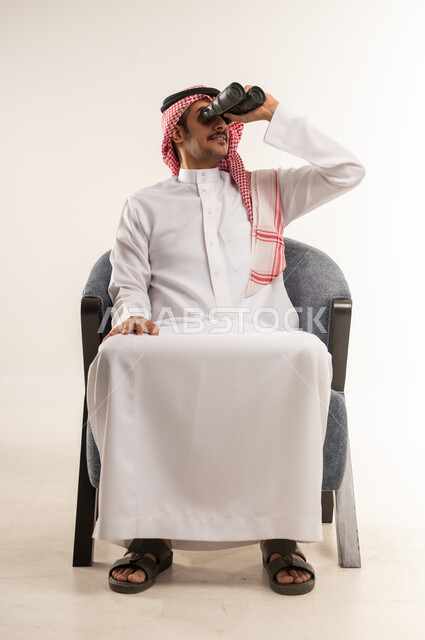 استخدام المنظار ثنائي العينين لمشاهدة الأشياء البعيدة، التخطيط ووضع الأهداف المستقبلية، توضيح الرؤية وتقريبها، بورتريه لشاب عربي خليجي بحريني يرتدي الثوب التقليدي والشماغ يجلس على كرسي ويضع تلسكوب ذو العدسات المقربة على عينيه، الطول الكامل للجسم، خلفية بيضاء