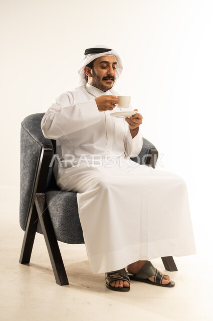الاسترخاء وتناول مشروبات دافئة، الاستمتاع بكوب من القهوة الساخنة الغنية، بورتريه لرجل عربي خليجي قطري يرتدي الثوب التقليدي والغترة يجلس على كرسي ويمسك بيده فنجان من مشروبه المفضل، محاولة الحصول على القليل من الراحة، خلفية بيضاء
