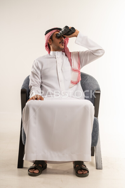 Clarifying and zooming vision, using binoculars to see distant objects, planning and setting future goals, portrait of a young Bahraini Gulf Arab man wearing traditional thobe and shemagh sitting on a chair and placing a telescope with telephoto lenses on his eyes, full body length, white background
