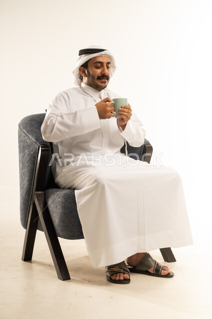 التلذذ بكوب من الشاي الساخن، محاولة الحصول على القليل من الراحة، الاسترخاء وتناول مشروبات دافئة، بورتريه لرجل عربي خليجي قطري يرتدي الثوب التقليدي والغترة يجلس على كرسي ويمسك بيده فنجان من مشروبه المفضل، خلفية بيضاء