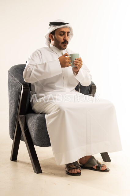 محاولة الحصول على القليل من الراحة، الاسترخاء وتناول مشروبات دافئة، بورتريه لرجل عربي خليجي قطري يرتدي الثوب التقليدي والغترة يجلس على كرسي ويمسك بيده فنجان من مشروبه المفضل، التلذذ بكوب من الشاي الساخن، خلفية بيضاء