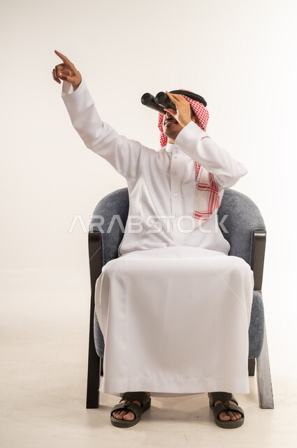 Pointing to a distant place, using binoculars to clarify and zoom in, planning and setting future goals, portrait of a young Bahraini Gulf Arab man wearing traditional thobe and shemagh sitting on a chair and placing a telescope with telephoto lenses on his eyes, full body length, white background