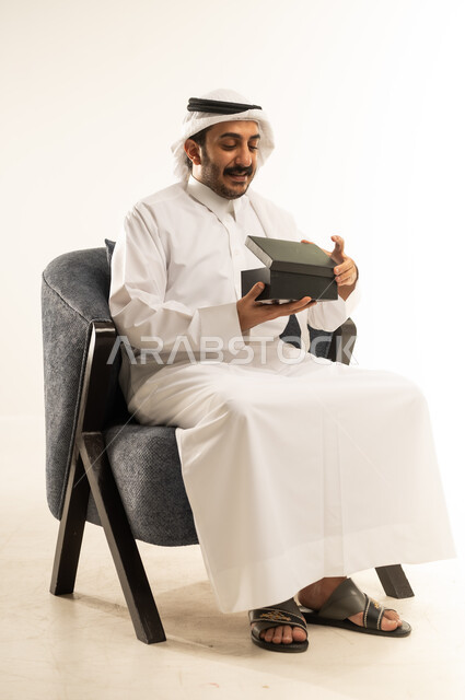 مفهوم المفاجأة والإهداء، إيماءات السعادة والسرور بالحصول على هدية ثمينة، فن انتقاء الهدايا بعناية، بورتريه لرجل عربي خليجي قطري يرتدي الثوب التقليدي والغترة يجلس على كرسي ويقوم بفتح علبة باللون الأسود، خلفية بيضاء