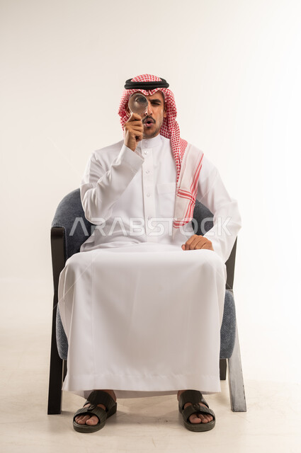 عمليات البحث والتدقيق، تكبير وتوضيح الرؤية باستخدام العدسة المكبرة، مضاعفة الأحجام ومحاولة إيجاد الأشياء الصغيرة، بورتريه لشاب عربي خليجي بحريني يرتدي الثوب التقليدي والشماغ يجلس على كرسي يحاول اكتشاف شيء ما بإيماءات الاندماج، الطول الكامل للجسم، خلفية بيضاء