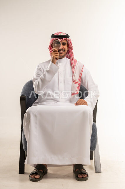 Zooming in and out using a magnifying glass, searching and scrutinizing, doubling sizes and trying to find small things, portrait of a young Arab Gulf Bahraini man wearing a traditional thobe and shemagh sitting on a chair trying to discover something with gestures of integration, full body length, white background